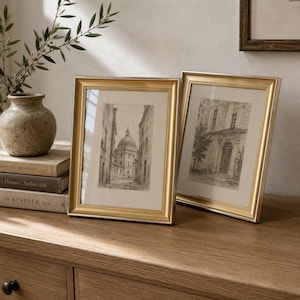May include: Two rectangular picture frames with metallic gold finishes and off-white matting. Each frame contains a black and white architectural sketch of a building. The frames are positioned upright, side by side, on a wooden surface.