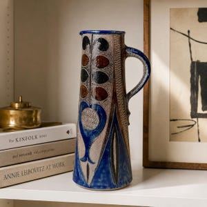 May include: A tall ceramic pitcher featuring a textured beige body with a vertical pattern of brown and black leaf-like shapes. The base and handle are finished in a deep blue glaze, with a prominent blue heart-shaped motif centered on the front.