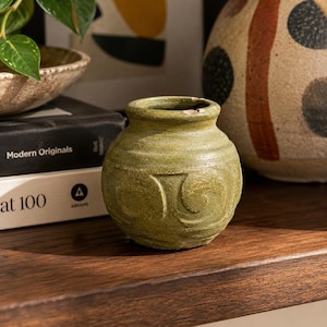 May include: A small, round, olive-green ceramic vase with a textured surface and a carved, swirling pattern around its centre. The vase features a short, thick neck with a slightly chipped rim, showcasing a matte, earthy finish and a rounded base.