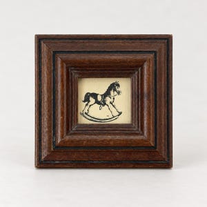 May include: A square, dark brown wooden frame containing a small, cream-colored print of a black rocking horse. The frame features a stepped, beveled design with dark recessed lines, surrounding the centered illustration of the toy horse.