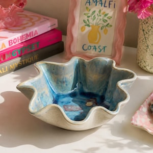 May include: A ceramic bowl with a wavy, scalloped edge. The bowl has a light blue and white glaze with darker blue accents in the center. Other items include books, a pink framed print, and a vase with pink flowers.