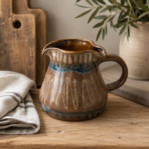Pode incluir: Um jarro de cerâmica com um esmalte castanho salpicado e padrões decorativos pintados em azul e creme. O recipiente tem um corpo largo e arredondado, uma pega curva no lado direito e um bico vertedor distinto no rebordo superior.