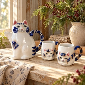 Puede incluir: Una tetera de cerámica blanca con forma de gato sentado con rayas azules, nariz rosa y asa en forma de cola curva, acompañada de dos tazas de cerámica blanca a juego que presentan un diseño de cara de gato en relieve con rayas azules.
