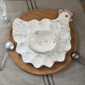 Puede incluir: Una fuente de cerámica blanca para servir huevos con forma de gallina, con un cuenco central y hendiduras en forma de huevo. La fuente tiene un estampado floral rojo y está sobre una bandeja de madera. Dos cucharas de plata a cada lado.
