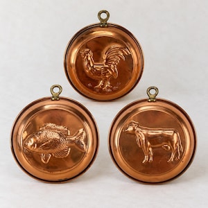 May include: Three circular copper molds featuring embossed animal designs. The top mold displays a rooster, the bottom left mold shows a fish, and the bottom right mold depicts a cow. Each mold includes a small metal hanging loop at the top.