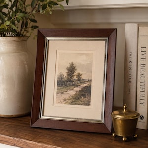 May include: A framed landscape print with a dark brown wooden frame and a silver inner border. The print depicts a dirt road, trees, and a fence. The frame is displayed on a wooden shelf with books and a decorative brass container.