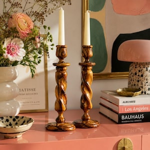 May include: Two tall, turned wooden candle holders with a dark brown, swirling grain pattern. Each holder features a spiraled central column, a circular base, and a top cup holding a single, plain, off-white tapered candle.