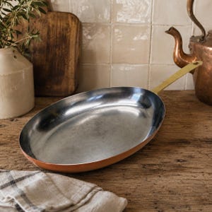 May include: An oval-shaped frying pan featuring a polished metallic interior surface, a warm copper-toned exterior, and a long, slender brass handle extending from the side, designed for stovetop cooking and culinary preparation in a kitchen.