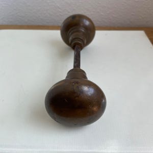 May include: Antique bronze door knob with a round, bulbous shape. The knob has a dark, aged patina, showing signs of wear. The connecting rod is a darker, contrasting metal. This vintage hardware adds a touch of classic charm.