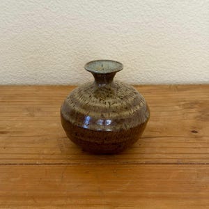 May include: A small, round ceramic vase with a narrow neck and a textured surface. The vase is primarily brown with lighter brown and tan horizontal bands. The rim of the vase is a muted green color. The vase sits on a wooden surface.