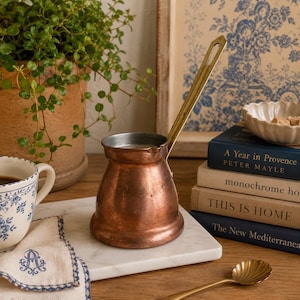 May include: A copper coffee pot with a long brass handle sits on a white marble surface. A white and blue floral teacup and a matching napkin are in the foreground. A small potted plant and a stack of books are in the background.