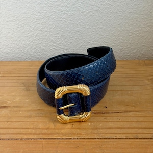 May include: A coiled navy blue belt with a textured snakeskin pattern. The belt features a large, square gold-colored buckle with a textured design. The belt is displayed on a wooden surface.