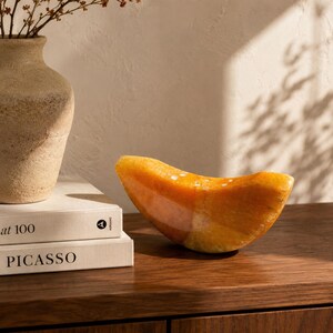May include: A decorative, abstract sculpture in shades of orange and yellow, resembling a curved slice. The sculpture sits on a dark wooden surface, next to a beige vase with dried branches and a stack of books.