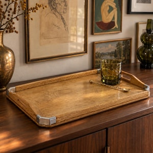 May include: A rectangular wooden serving tray featuring a natural wood grain finish, integrated side handles, and polished silver-tone metal corner accents. A textured olive green glass tumbler and a thin gold-tone metal stirring spoon rest on top.