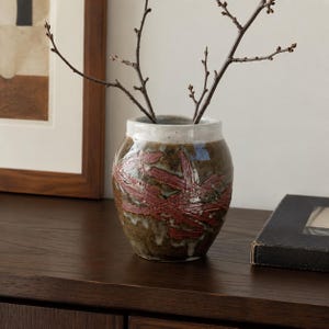 May include: A ceramic vase with a rounded body, featuring a mottled dark brown and white glaze with textured, raised red brushstroke patterns. The vase has a wide, open mouth and contains a thin, bare, dark brown branch with small buds.