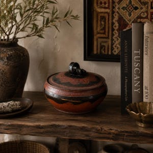 May include: A handcrafted ceramic lidded jar with a dark red and black design. The jar has a round shape and a black handle on the lid. It sits on a rustic wooden shelf, alongside books and decorative items.