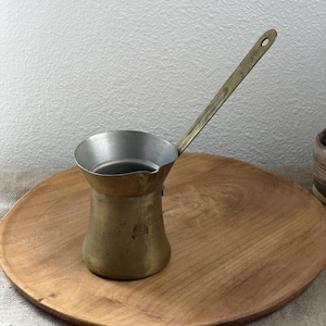 May include: A vintage brass coffee pot, also known as a cezve, with a long handle and a silver-colored interior. The pot is on a wooden serving tray. The handle has a small hole at the end.