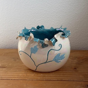 Puede incluir: Un cuenco de cerámica con acabado nacarado y borde con volantes. El interior es de color verde azulado intenso y el exterior presenta un delicado diseño de hojas azules. El borde tiene un ribete dorado.