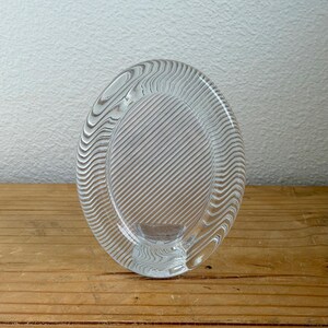 Peut inclure: Cadre photo ovale en verre transparent avec une bordure texturée ondulée et des lignes diagonales. Le cadre est conçu pour tenir debout et est en verre transparent. Le cadre est sur une surface en bois.