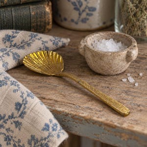 May include: A gold-toned metal spoon with a bowl shaped like a seashell, featuring a textured handle. Beside the spoon sits a small, round, speckled beige stone bowl filled with coarse white salt crystals, resting on a wooden surface.