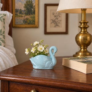 Peut inclure: Un cache-pot en céramique bleu clair en forme de cygne, doté de détails de plumes moulés sur le corps et d'un long cou incurvé. Le cygne contient un petit bouquet de fleurs blanches au cœur jaune, formant un arrangement décoratif délicat.