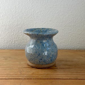 May include: A small, speckled blue ceramic vase with a wide, ruffled rim. The vase has a rounded body and sits on a circular, unglazed base. The blue glaze has darker blue and brown speckles, giving it a rustic appearance. The vase is displayed on a wooden surface.