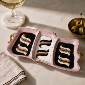 May include: A pink, three-section serving tray with gold trim and decorative accents. Each section features a black or white background with a gold and black stylized design. The tray is designed for serving appetizers or snacks.