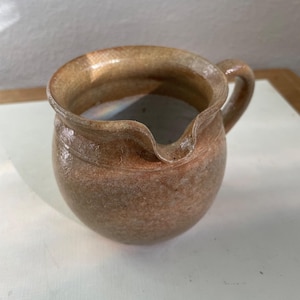 May include: A small, round, ceramic pitcher with a handle and a spout. The pitcher is a mottled brown color with a lighter rim and handle. The pitcher is likely handmade and has a rustic appearance.
