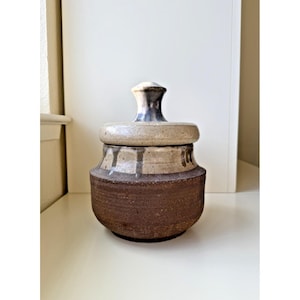 Könnte beinhalten: Ein handgefertigter Keramiktopf mit Deckel. Der Topf hat einen dunkelbraunen Boden, einen beigefarbenen Mittelteil mit vertikalen Akzenten und einen hellbraunen Deckel. Der Deckel hat einen dunkelgrauen Griff. Der Topf ist ein dekorativer Aufbewahrungsbehälter.