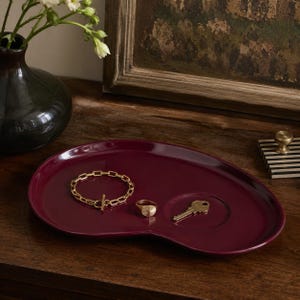 May include: A deep burgundy, kidney-shaped ceramic tray with a smooth, glossy finish. Resting on the surface are a gold-toned chain link bracelet with a toggle clasp, a gold-toned textured ring, and a single brass-colored metal key.