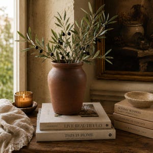 Könnte beinhalten: Eine rustikale Vase aus Terrakotta mit Olivenzweigen und schwarzen Oliven. Die Vase steht auf einem Stapel Bücher, darunter "Live Beautifully" und "This is Home". Eine brennende Kerze in einem Glas und eine kleine Schale ergänzen die Szene.