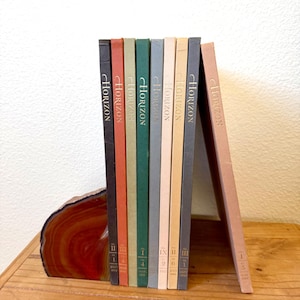May include: A collection of vintage "Horizon" magazines with various colored spines, including black, orange, green, and gray. The magazines are held upright by a decorative agate bookend. The magazines are on a wooden surface.