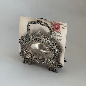 May include: An antique silver-colored basket with an ornate floral design. The basket has a handle and a decorative front with a curved shape. A vintage postcard with a red stamp is inside the basket.