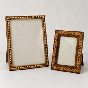 May include: Two rectangular wooden picture frames with glass inserts. The larger frame features a light-colored diamond inlay pattern along the border. The smaller frame displays a darker wood grain with a geometric square pattern border.