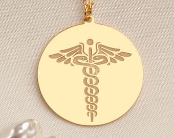 May include: A gold-colored pendant necklace featuring a circular charm with a medical symbol. The charm is suspended from a delicate gold chain. The background is a neutral beige color, with several white, irregularly shaped pearls scattered around the necklace.