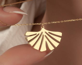 May include: A gold-colored necklace with a fan-shaped pendant. The pendant has a series of vertical lines radiating from a central point. The necklace is being held by a person, and the chain is delicate.
