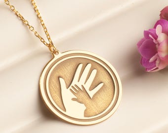 May include: A gold-colored pendant necklace featuring a circular charm with a hand-in-hand design. The necklace is displayed with a sprig of pink flowers. The chain and pendant are made of a shiny, reflective metal.