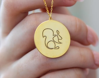 May include: Gold-colored pendant necklace featuring a circular charm with a squirrel design. The squirrel is depicted in a minimalist line art style, holding a nut. The necklace has a delicate chain and is held up by a hand.