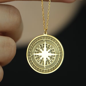 May include: Gold-colored compass pendant necklace. The pendant is round with a detailed compass design. The necklace is held up by a hand, showcasing the delicate chain and pendant. The compass design features a star in the center.