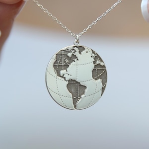 Peut inclure: Un collier avec un pendentif en forme de globe argenté. Le pendentif représente la Terre, avec les continents et les océans. Le collier est tenu par une personne aux ongles manucurés.