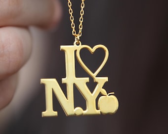 May include: Gold-colored necklace pendant spelling "I Love NY" with a heart and an apple. The pendant hangs from a delicate chain. The necklace is held up by a person's hand.