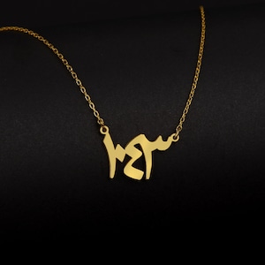 Könnte beinhalten: Eine goldene Halskette mit einer zarten Kette und einem Anhänger mit einem Namen in arabischer Schrift.