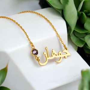 May include: Gold necklace with a name written in Arabic script and a small purple gemstone.