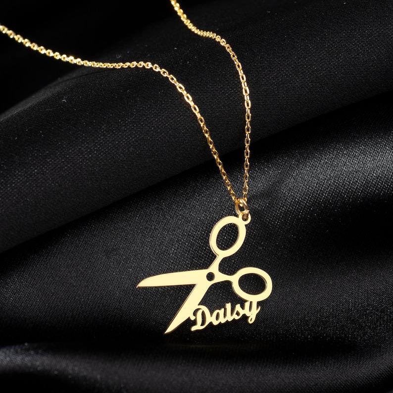 May include: Gold necklace with a pendant featuring a pair of scissors and the name "Daisy" written in cursive.