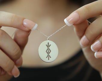 May include: A silver pendant necklace featuring a circular charm with an engraved symbol. The pendant hangs from a delicate chain. The symbol is a stylized design with vertical and angled lines. The necklace is held up by a person.