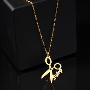 May include: Gold necklace with a pendant shaped like a pair of scissors with the name "Daisy" written on the side.
