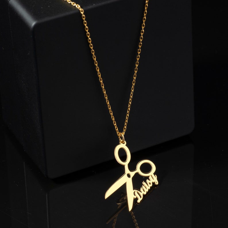 May include: Gold necklace with a pendant shaped like a pair of scissors with the name "Daisy" written on the side.