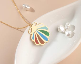 May include: A gold-colored necklace with a seashell pendant featuring colorful enamel stripes of red, brown, blue, green, and light blue. The necklace is displayed on a white dish with several white pearls.