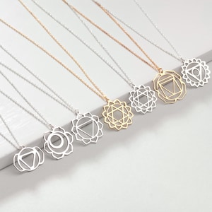 Könnte beinhalten: Sieben silberne und goldene Halsketten mit verschiedenen Chakra-Symbolen. Jede Halskette hat eine zarte Kette und einen Anhänger mit einem einzigartigen geometrischen Design.
