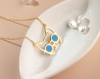 May include: A gold-colored necklace featuring a cat-shaped pendant with blue enamel accents. The necklace is displayed on a white, oval-shaped dish, with several white pearls nearby.